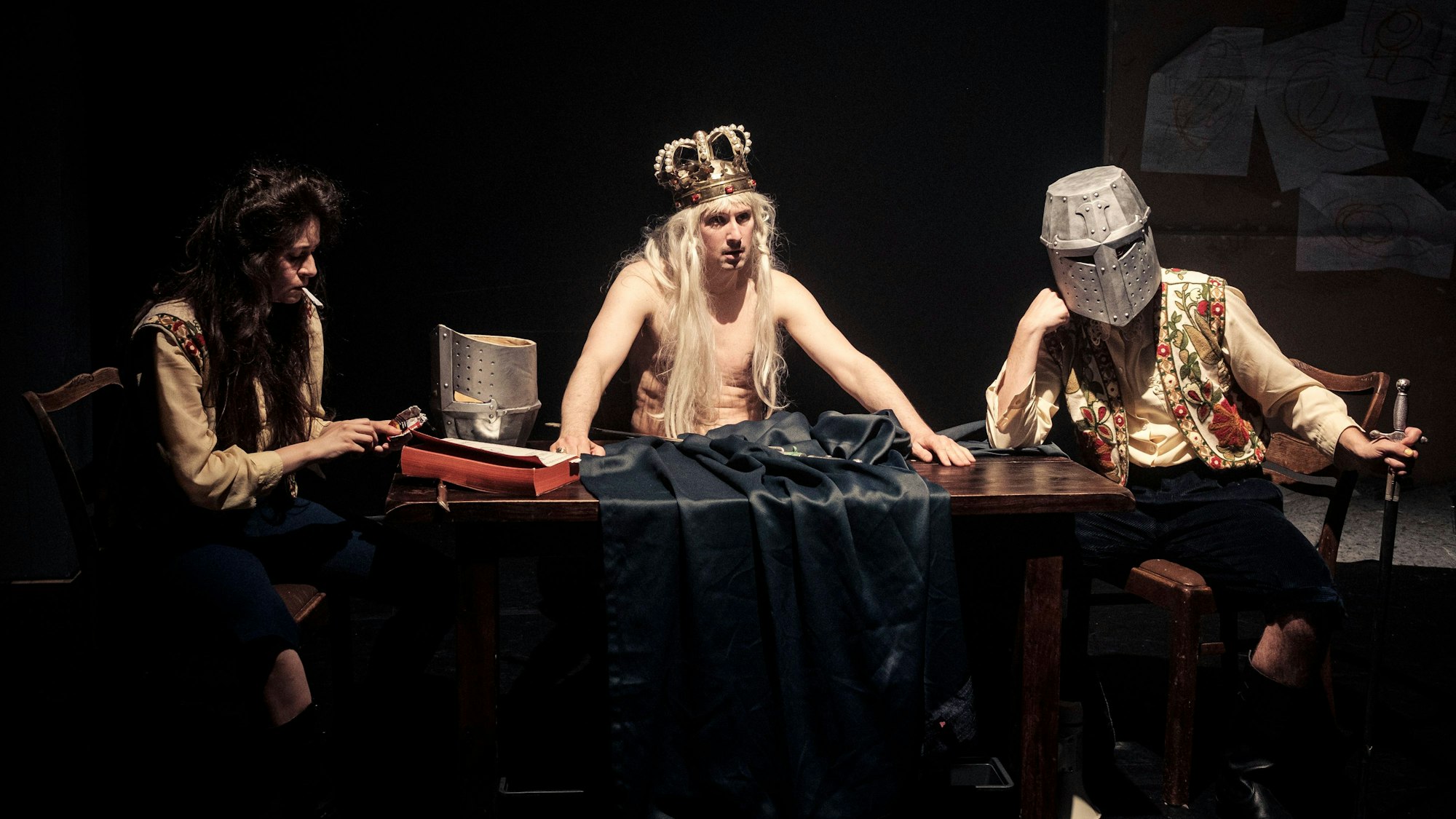 König Ubu sitzt mit freiem Oberkörper und pompöser Krone an einem Tisch, rechts und links neben ihm seine zwei Gefolgsleute mit Zigarette und Ritterhelm.