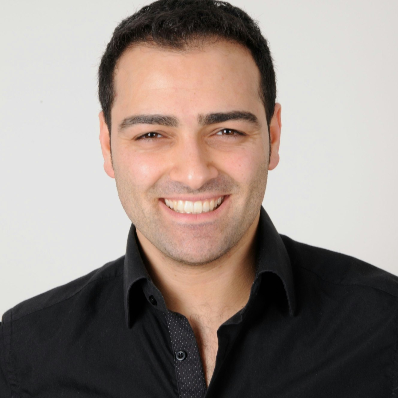 Numan Sarrac ist im Porträt zu sehen, er lacht in die Kamera. Er hat kurzes dunkles Haar und trägt ein schwarzes Hemd.