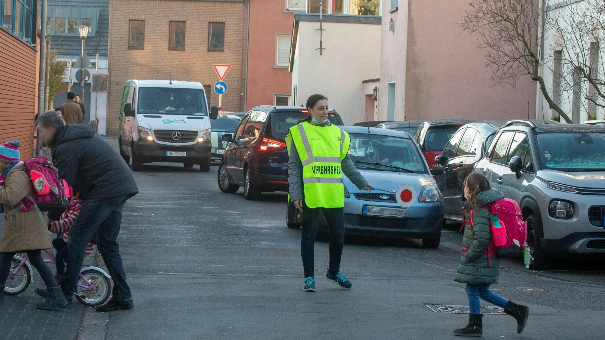 Eine Verkehrshelferin in gelber Warnweste lots Schulkinder und einen Vater über die Straße. Im Hintergrund sind Fahrzeuge zu sehen, die sich in der Straße Schulpfad selbst blockieren