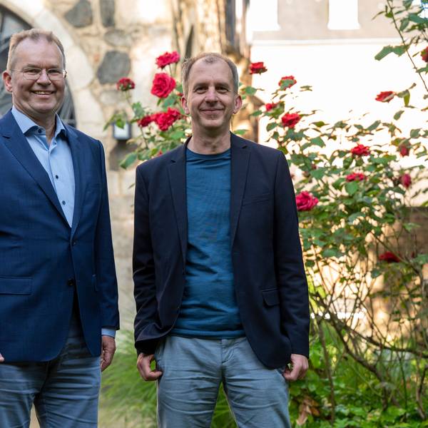 Zwei Männer mittleren Alters in blauen Sakkos vor dem Garten einer Kirche