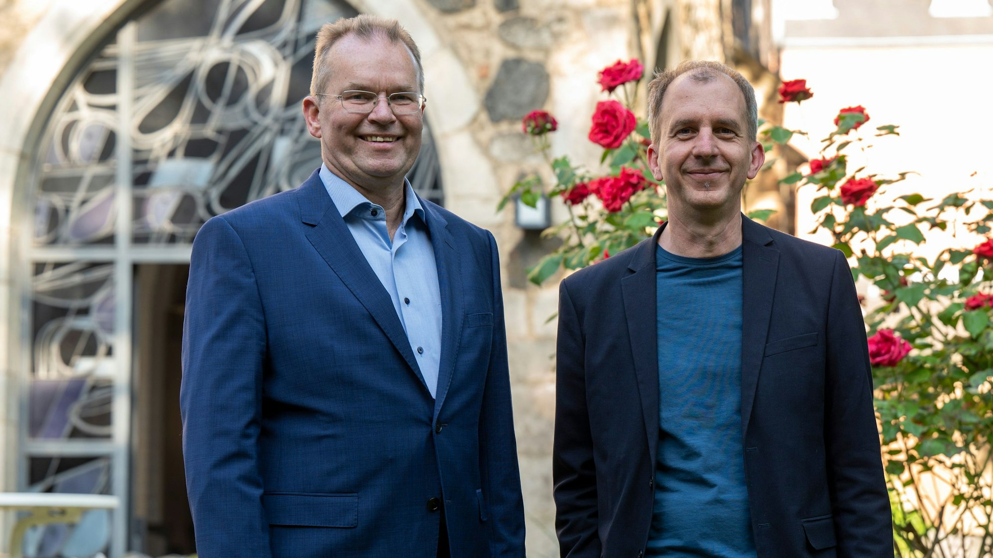 Zwei Männer mittleren Alters in blauen Sakkos vor dem Garten einer Kirche