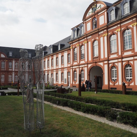 Zu sehen ist die Abtei Brauweiler mit ihrer weißen und terracotta-farbenen Fassade und einem Teil des Innenhofs.