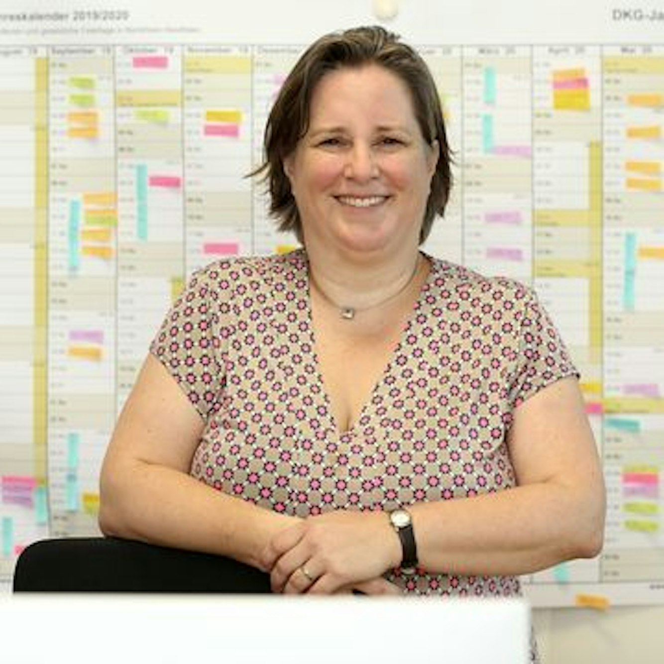 Eine Schulleiterin steht in einem bunten Kleid vor einem großen Wandkalender. Die Frau lacht sehr freundlich in die Kamera.