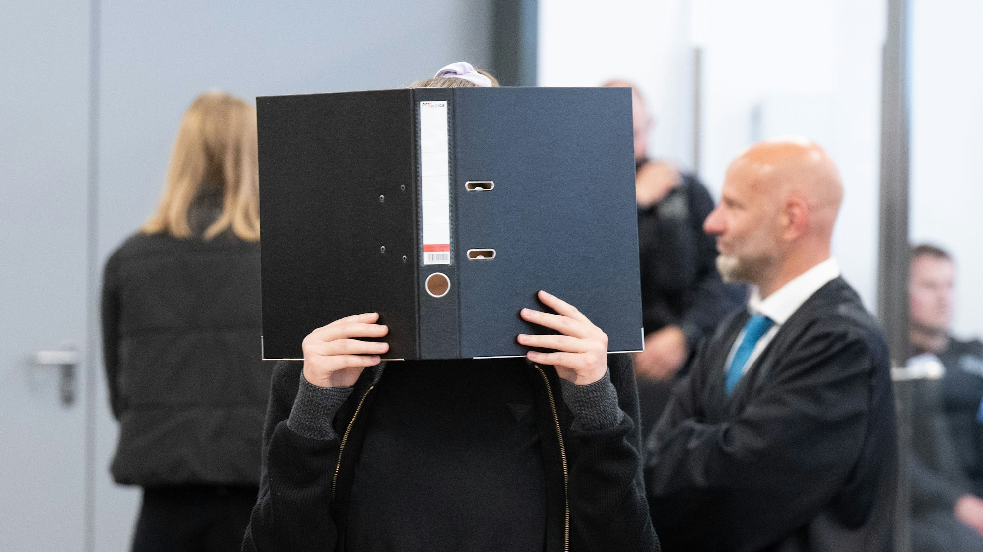 Die Angeklagte Lina E. steht bei der Fortsetzung des Prozesses im Oberlandesgericht (OLG) Dresden im Verhandlungssaal und hält einen Aktenordner vor ihr Gesicht.