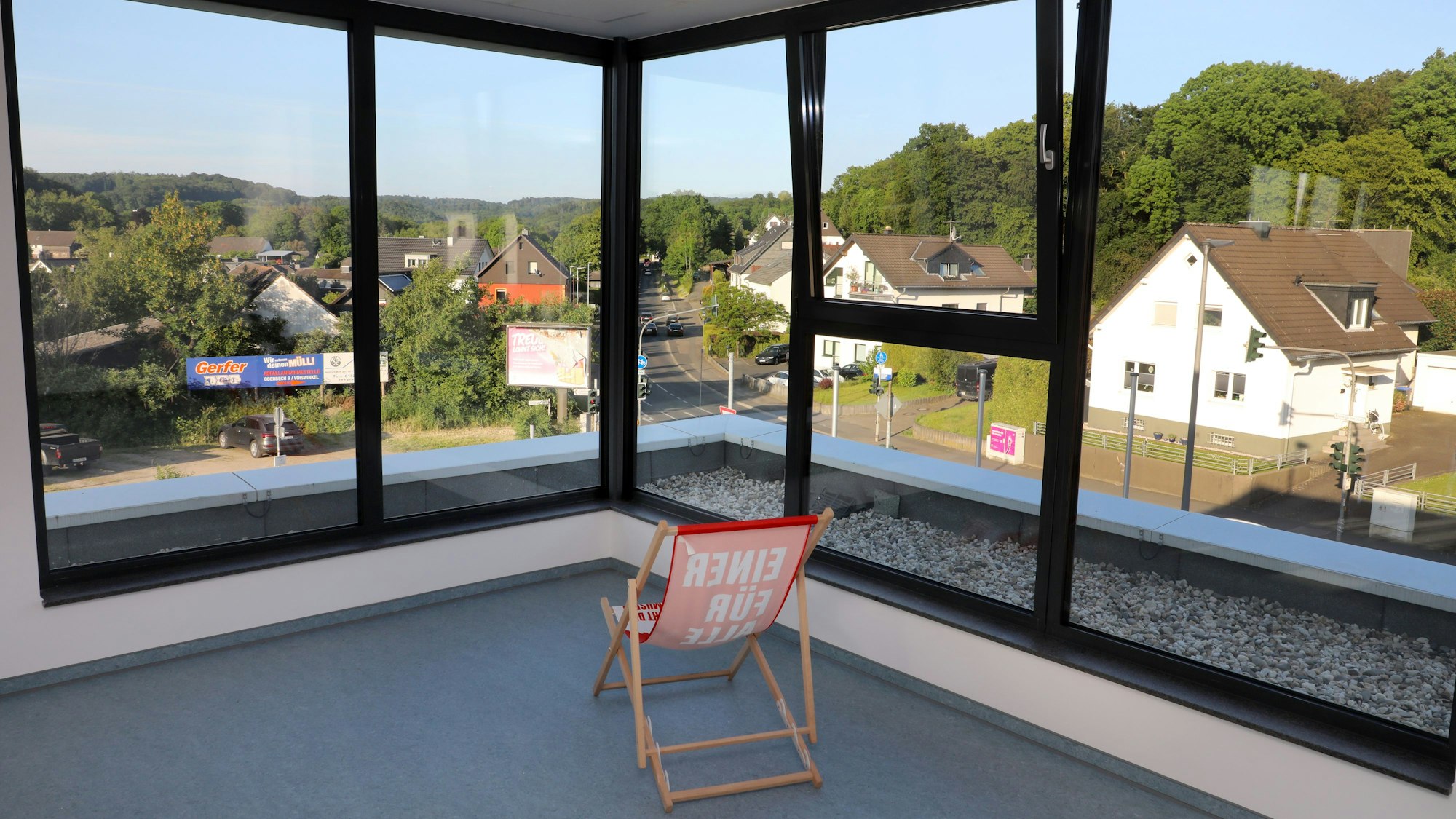Vor großen Panoramafenstern mit einem reizvollen Ausblick auf Huder, Straßen udn Bäume steht ein Liegestuhl in einem Raum.
