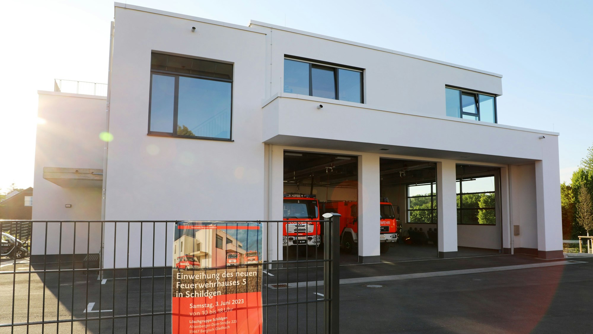 Im abendlicht steht ein Feuerwehrhaus mit geöffneten Fahrzeughallentoren. Am Zaun hängt ein Plakat, das auf den Tag der öffenen Tür am Samstag, 3. Juni, hinweist-