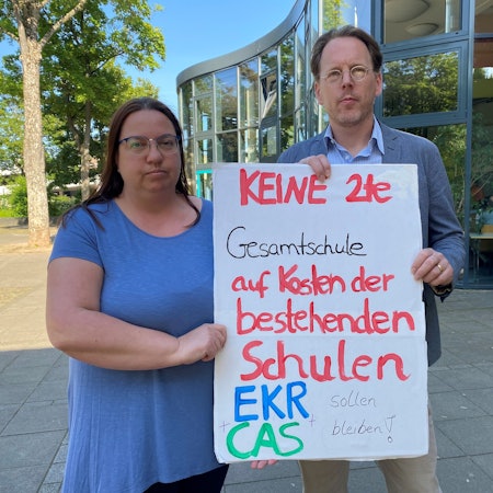 Es sind zwei Menschen zu sehen, die ein Schild hochhalten, auf dem steht: „Keine 2te Gesamtschule auf Kosten der bestehenden Schulen“.
