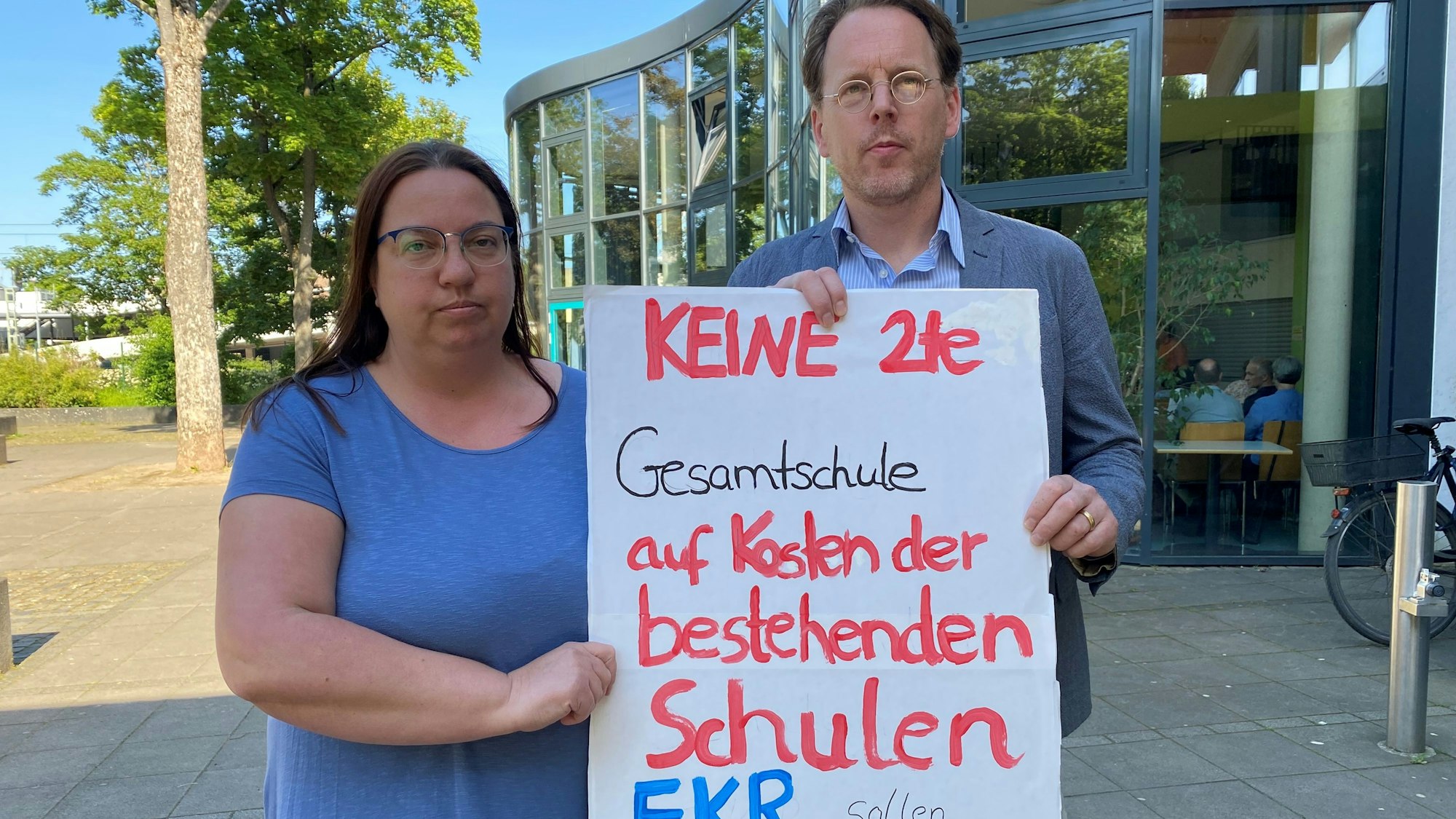 Es sind zwei Menschen zu sehen, die ein Schild hochhalten, auf dem steht: „Keine 2te Gesamtschule auf Kosten der bestehenden Schulen“.