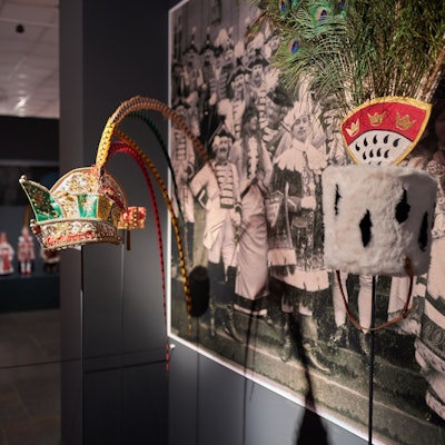 Zu sehen ist eine traditionelle Kopfbedeckung aus dem Kölner Karneval. Im Hintergrund sieht man ein Foto mit Menschen, die in traditioneller Karnevalskleidung feiern.