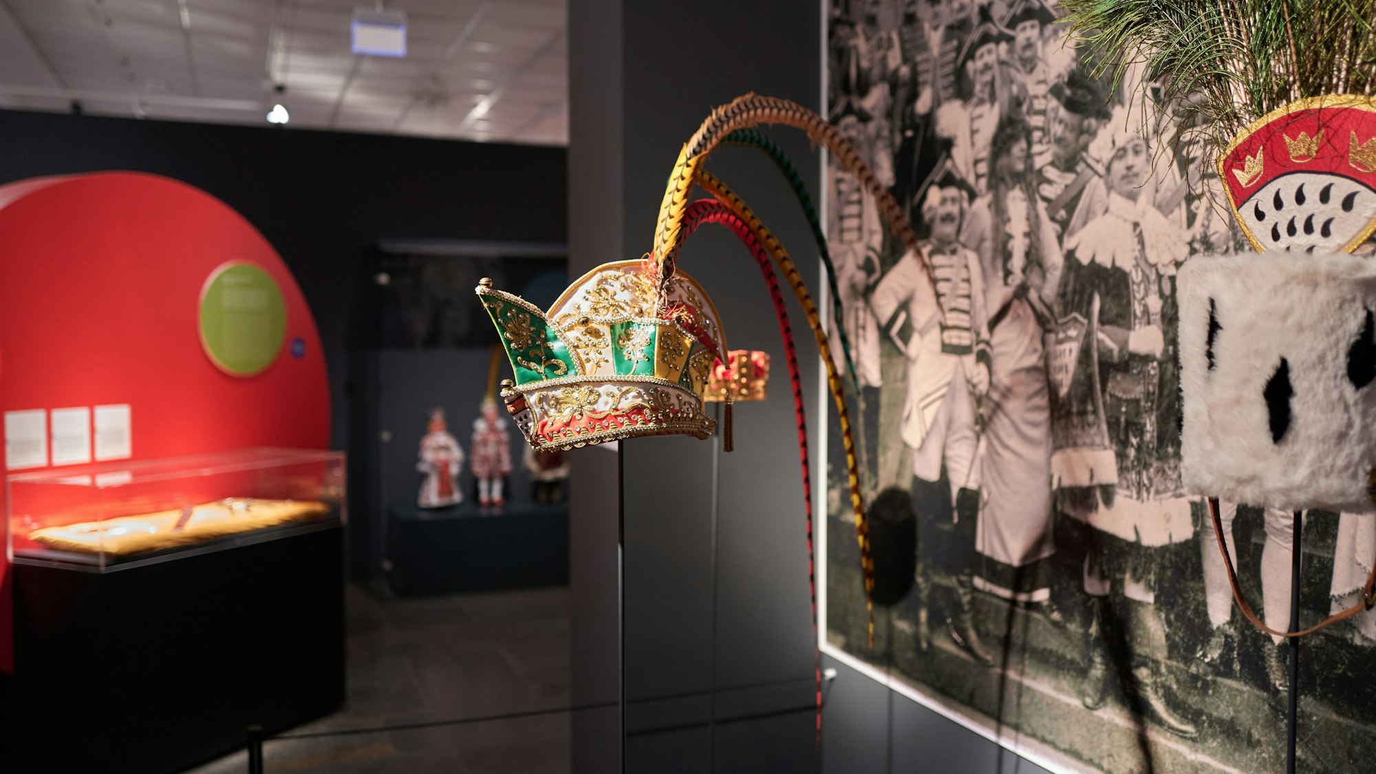 Zu sehen ist eine traditionelle Kopfbedeckung aus dem Kölner Karneval. Im Hintergrund sieht man ein Foto mit Menschen, die in traditioneller Karnevalskleidung feiern.