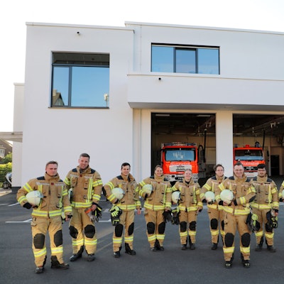 Feuerwehrleute stehen vor einem Feuerwehrhaus mit geöffneten Fahrzeughallentoren, in denen zwei große Einsatzfahrzeugezu sehen sind.