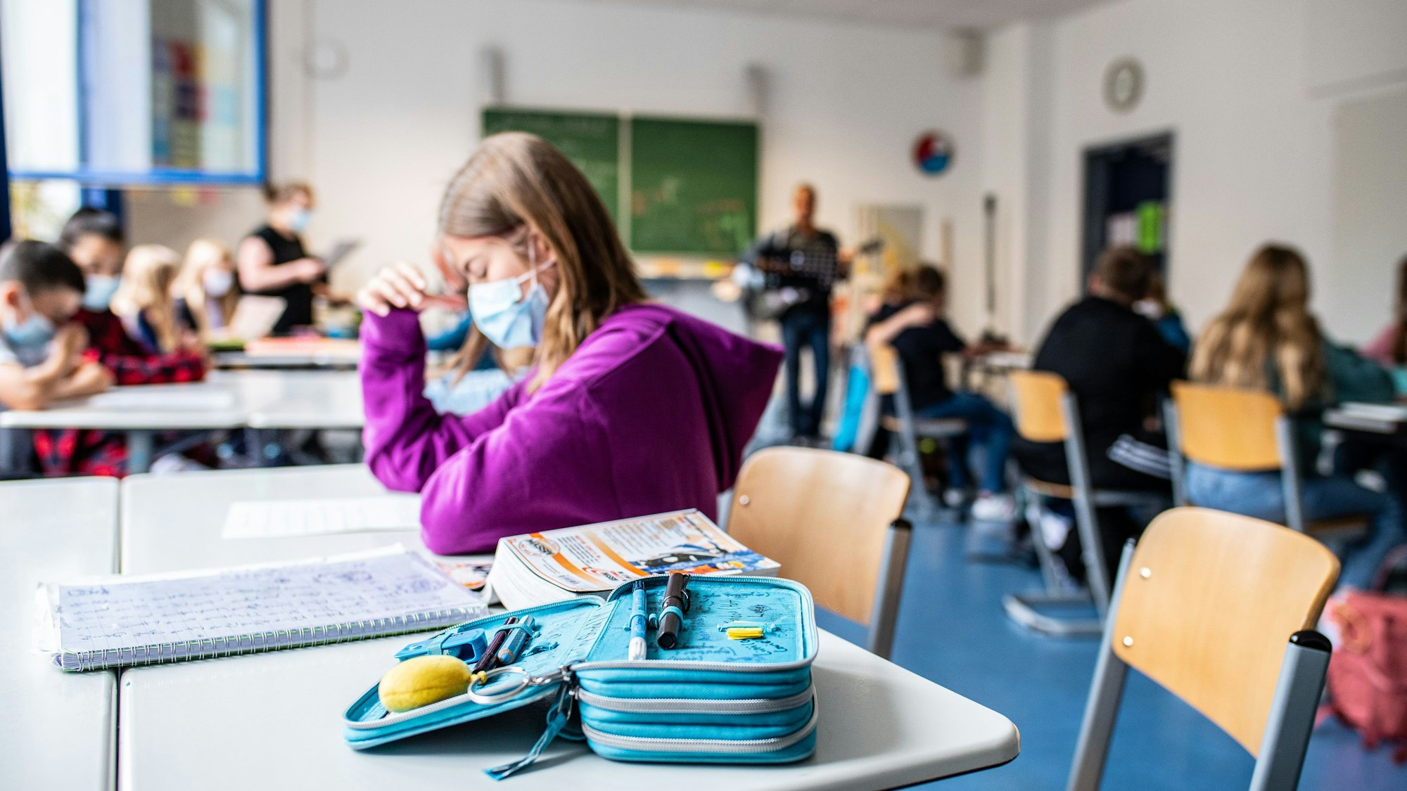 Eine Schülerin sitzt mit ihrer Maske neben ihrem Etui, das auf ihrem Tisch in der Klasse liegt.