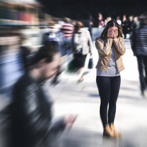 Eine junge Frau steht auf einer Straße und hält die Hände vors Gesicht, die Passanten um sie herum sind verschwommen, sie hat offenbar eine Panikattacke.