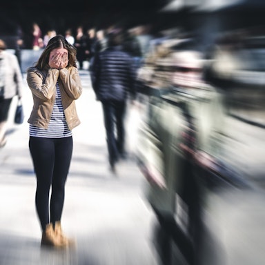 Eine junge Frau steht auf einer Straße und hält die Hände vors Gesicht, die Passanten um sie herum sind verschwommen, sie hat offenbar eine Panikattacke.