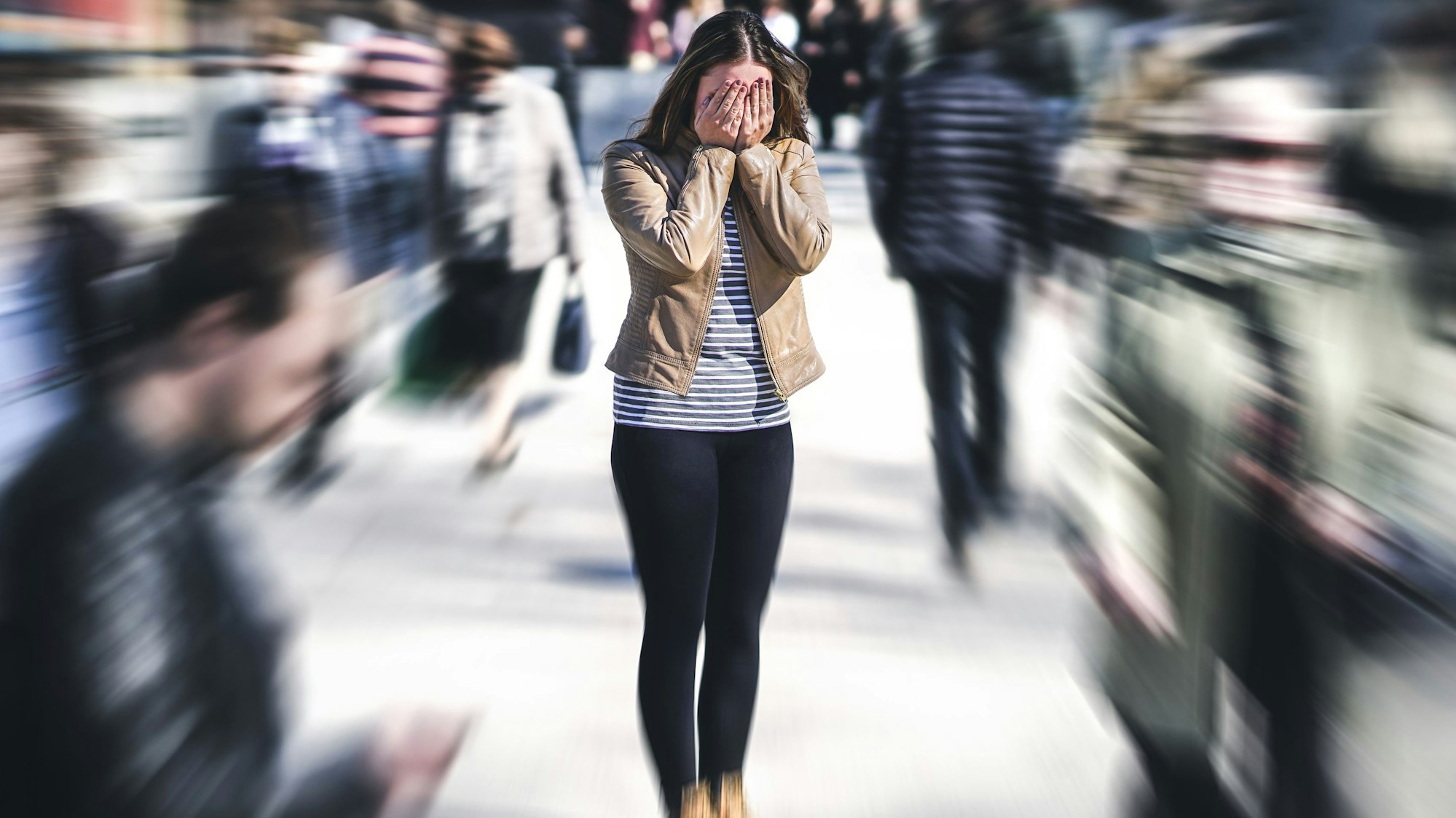 Eine junge Frau steht auf einer Straße und hält die Hände vors Gesicht, die Passanten um sie herum sind verschwommen, sie hat offenbar eine Panikattacke.