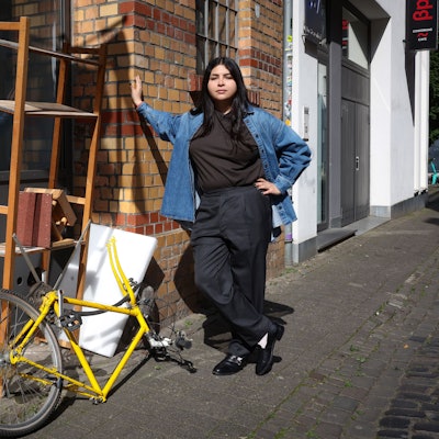 Die Künstlerin posiert neben einem kaputten Regal, einem Fahrrad ohne Vorderreifen und einem Vogelhäuschen.