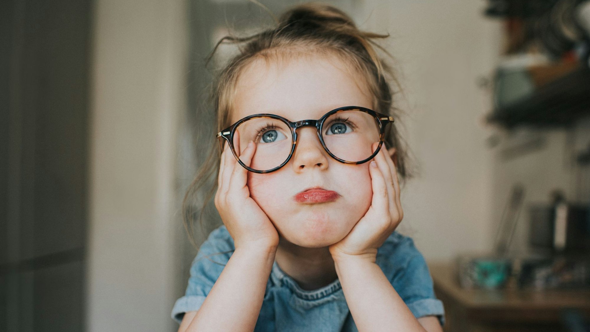 Ein kleines Mädchen mit einer großen, runden Brille hat sein Gesicht auf seine Hände gestützt und schaut sehr gelangweilt aus.