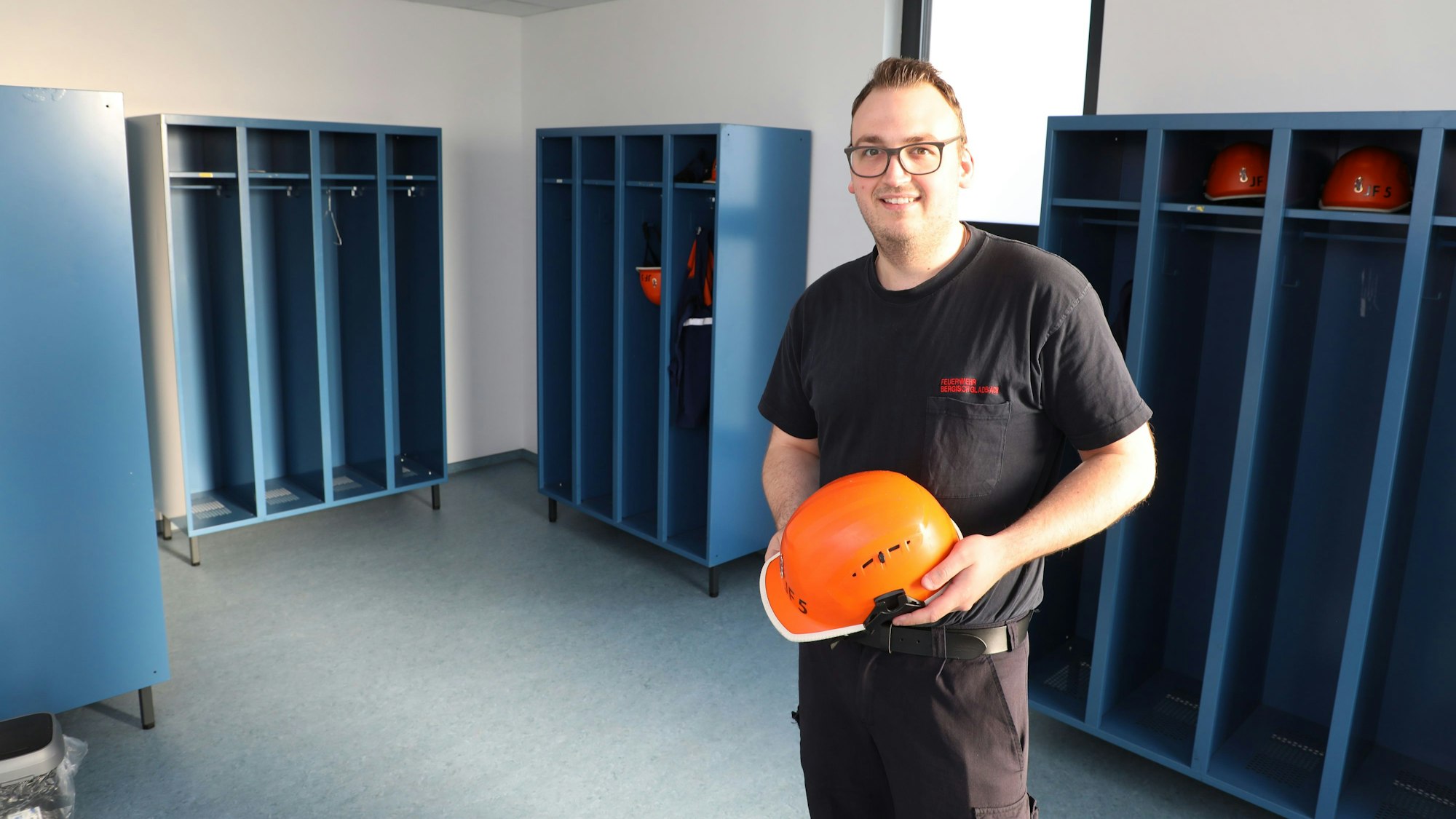 Jugendfeuerwehrwart Fabian Kaltenbach steht, einen Jugendfeuerwehrhelm haltend, in der neuen Umkleide samt Spinden der Jugendfeuerwehr Schildgen.