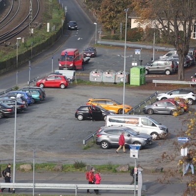 Parkende Autos stehen auf einer Schotterfläche. Links am Bildrand verläuft die rechtsrheinische Bahntrasse.