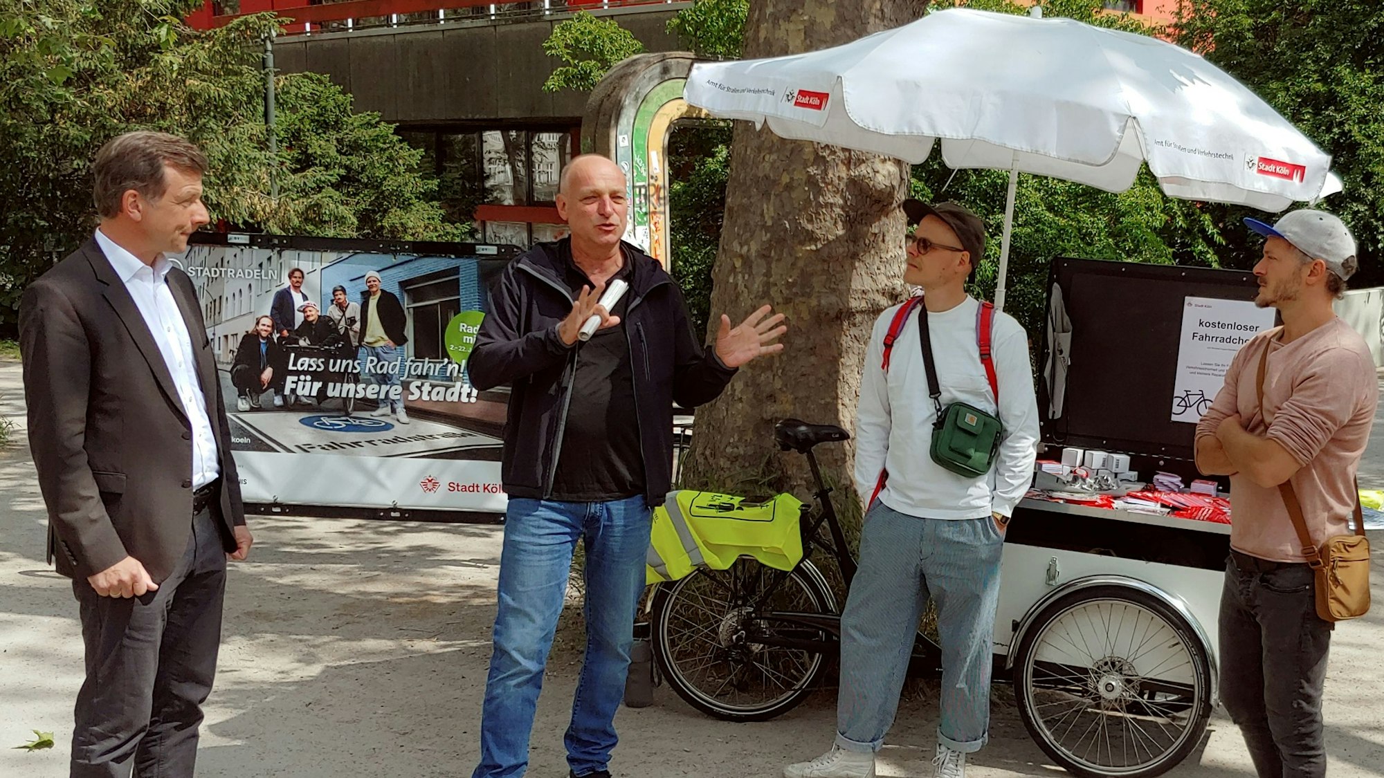 Die vier stehen um einen Lastenfahrrad-Stand mit Schirm.