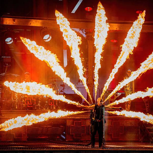 Rammstein-Sänger Till Lindemann steht bei einem Konzert auf der Bühne, er hat die Arme ausgebreitet und singt ins Mikrofon. Auf dem Rücken trägt er einen Pyrotechnik-Apparat, der Flammen zu allen Seiten schießen lässt.