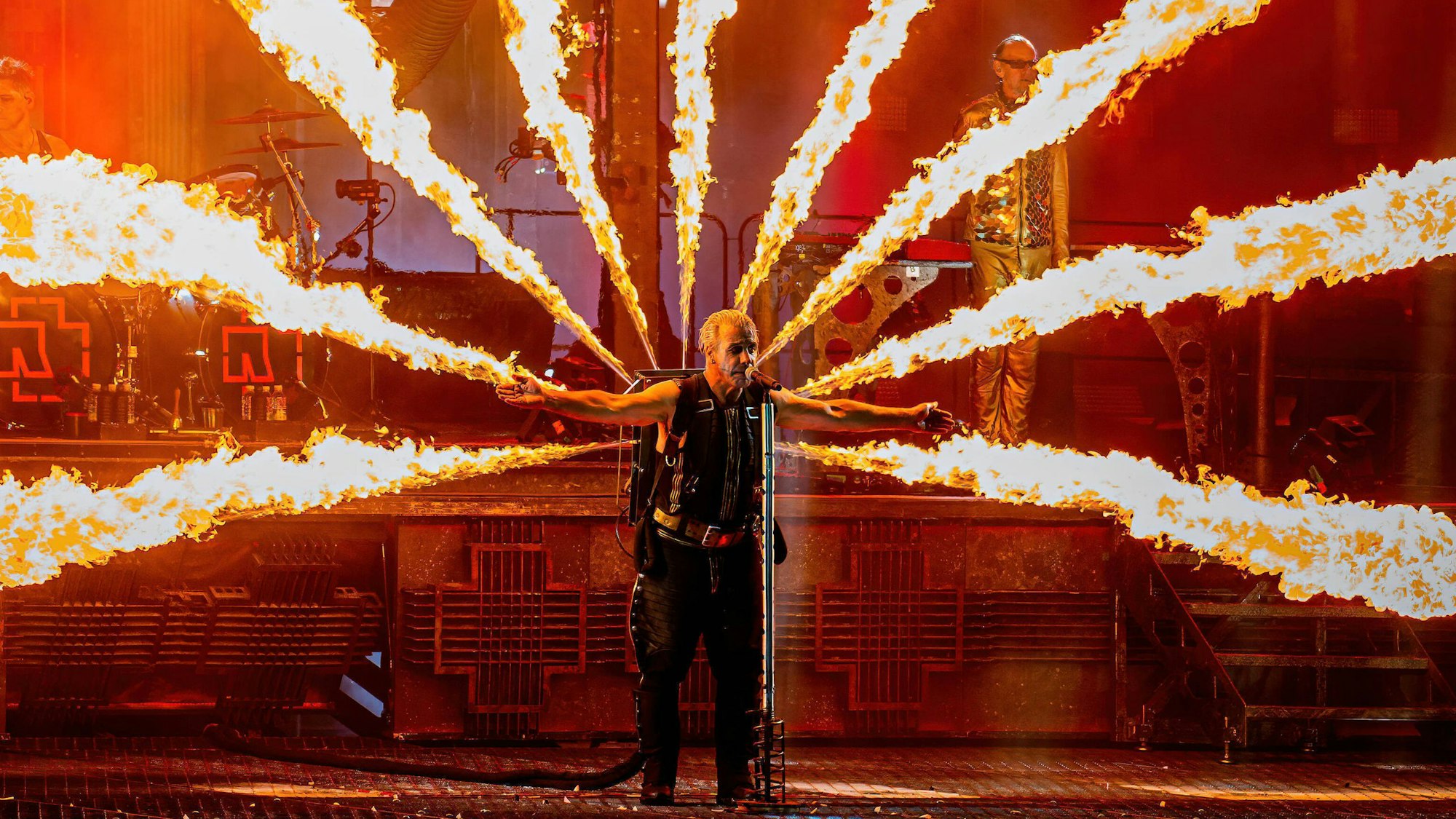 Rammstein-Sänger Till Lindemann steht bei einem Konzert auf der Bühne, er hat die Arme ausgebreitet und singt ins Mikrofon. Auf dem Rücken trägt er einen Pyrotechnik-Apparat, der Flammen zu allen Seiten schießen lässt.