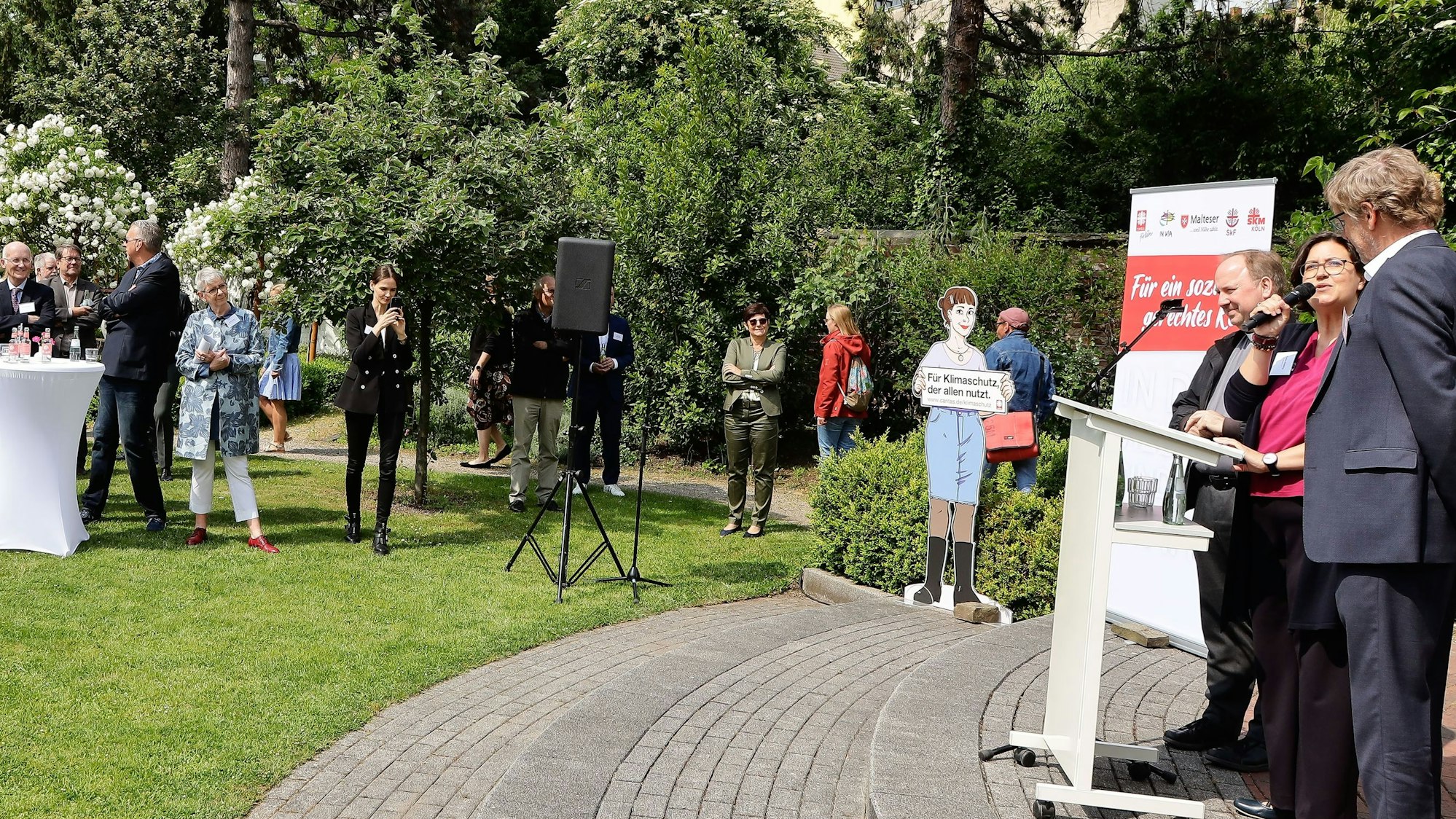 Im Garten aufgebautes Podium mit den Rednern bei sonnigem Wetter. Hinten und links im Bild das Publikum.