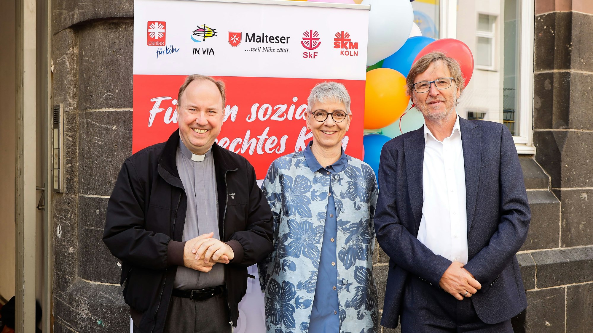 Die drei stehen vor einer Stellwand, die die Embleme verschiedener Wohlfahrtsverbände zeigt.