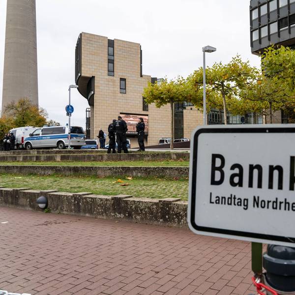 Das Schild mit Aufschrift ist zu sehen, dahinter Grünfläche, auf der einige Polizisten stehen und das Landtagsgebäude.