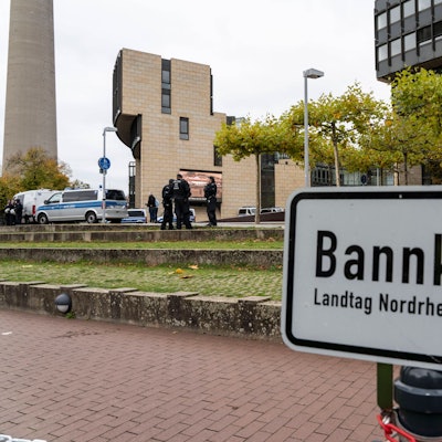 Das Schild mit Aufschrift ist zu sehen, dahinter Grünfläche, auf der einige Polizisten stehen und das Landtagsgebäude.