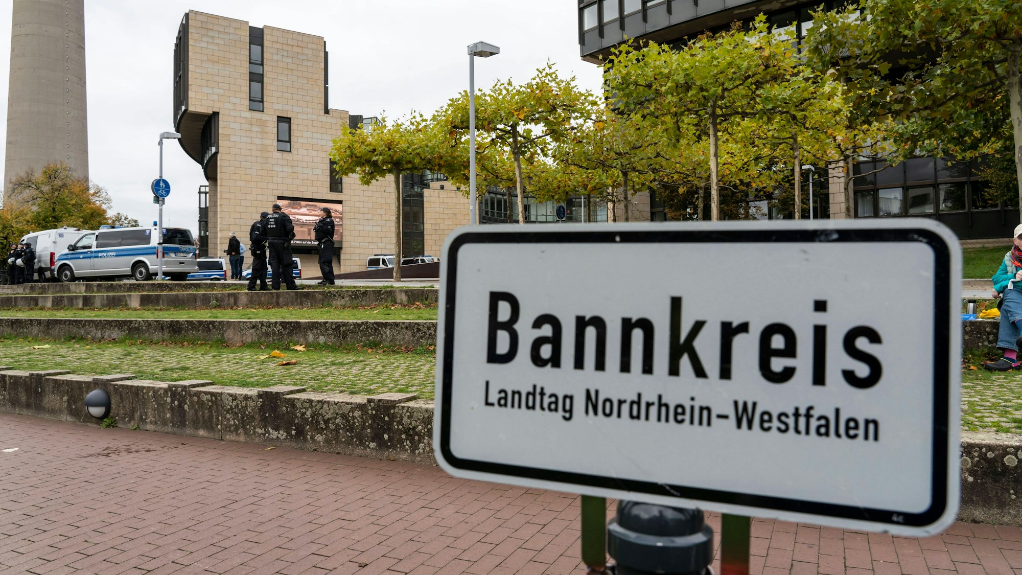 Das Schild mit Aufschrift ist zu sehen, dahinter Grünfläche, auf der einige Polizisten stehen und das Landtagsgebäude.