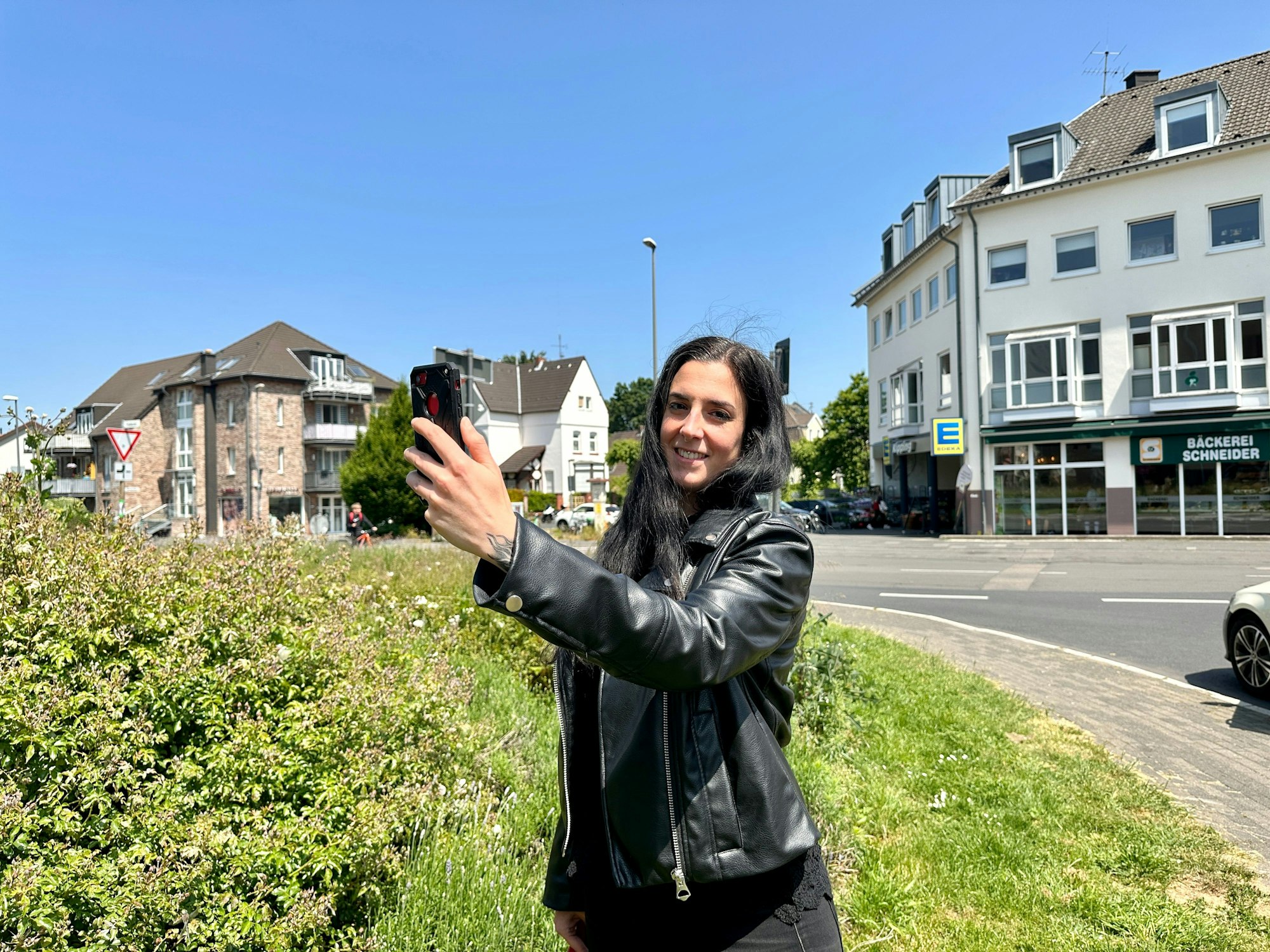 Eine junge Frau steht mit einem Smartphone in der Hand auf einer Grünfläche an einer Straße.