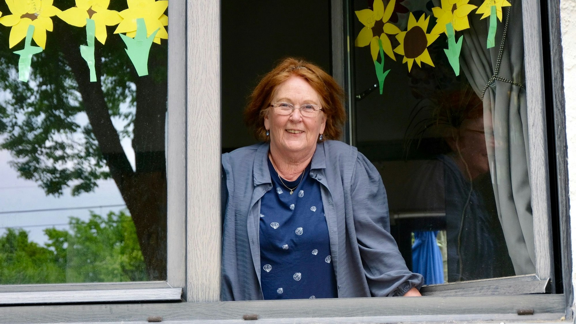 Mallat an einem mit Sonnenblumen verzierten Fenster. Sie schaut raus und in die Kamera
