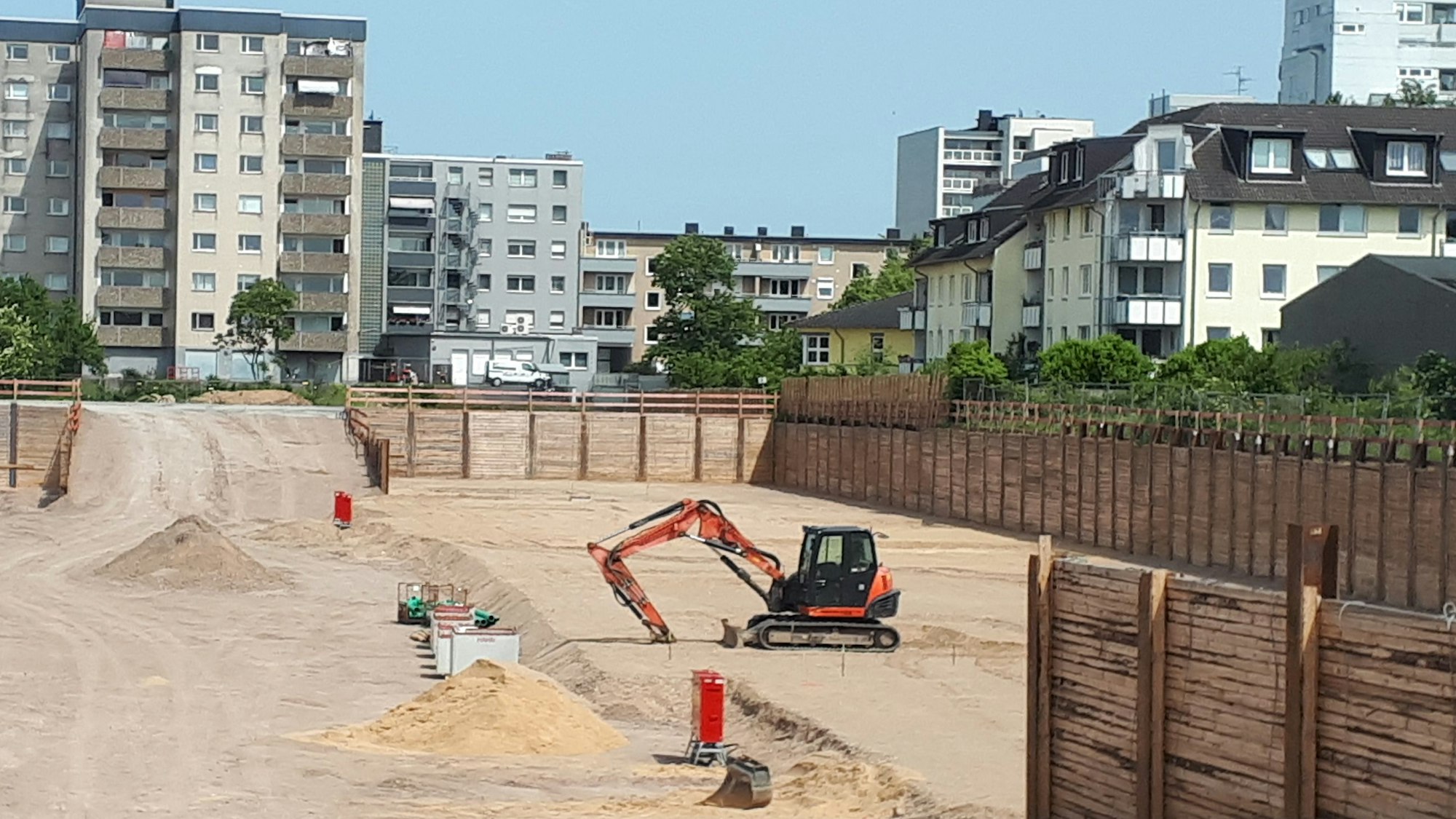 Im Hintergrund die Bebauung entlang der Neusser Straße entlang der Baugrube bei gutem Wetter. In der Baugrube steht ein kleiner Bagger.