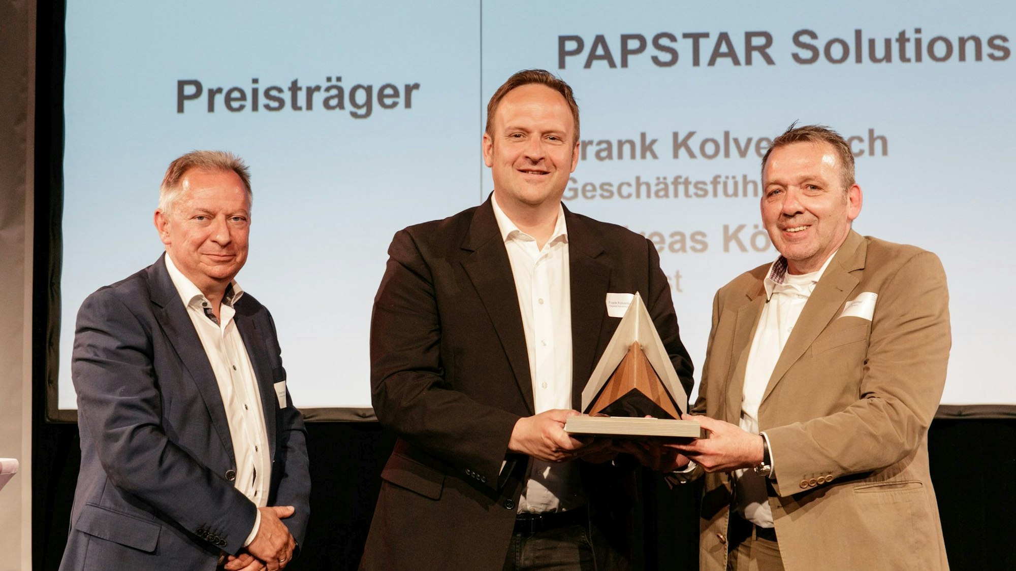 Drei Männer stehen vor einer Leinwand, zwei halten eine Skulptur für einen Wettbewerbssieg.