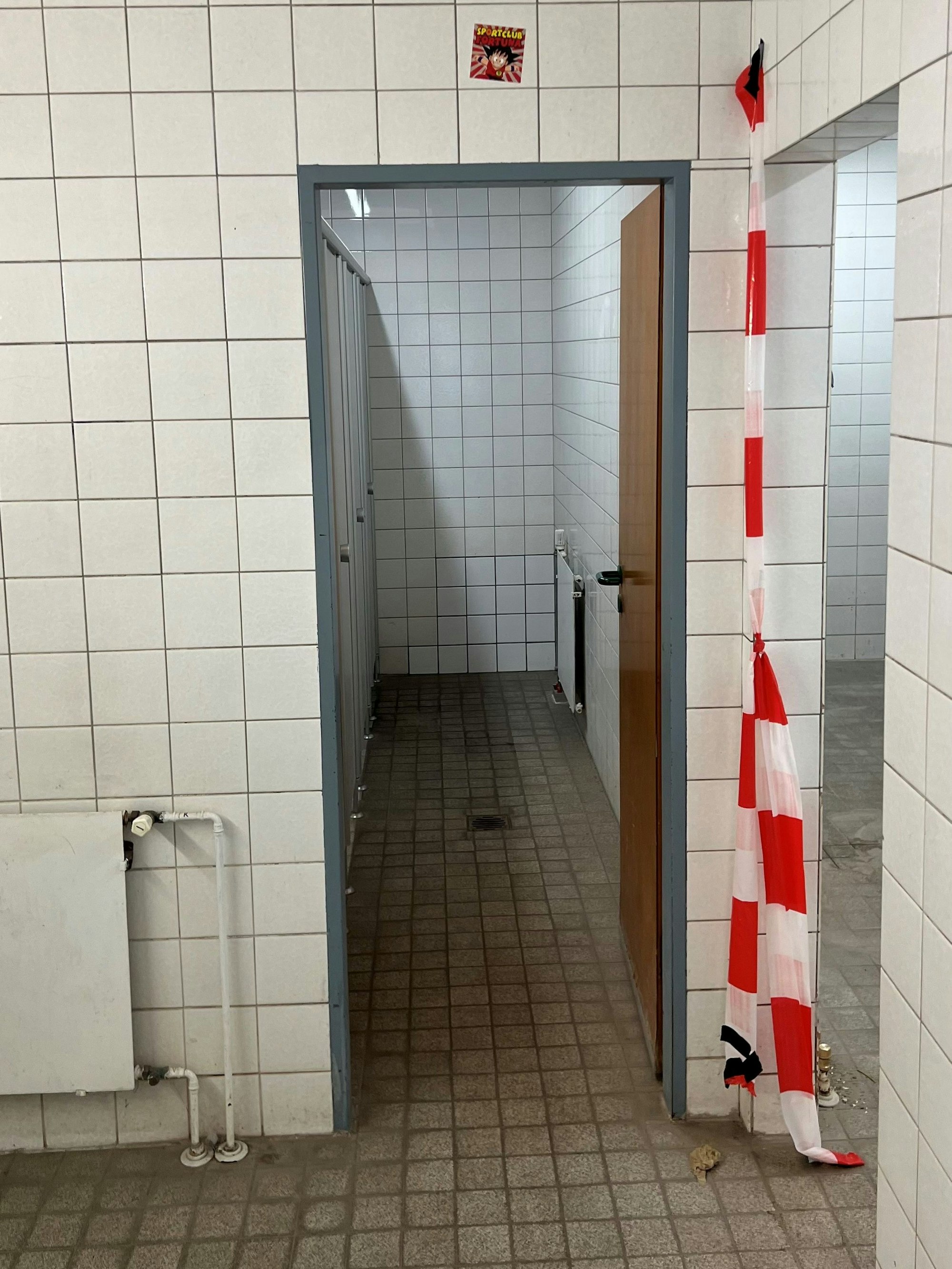 Alte Fliesen und Dreck ist zu sehen, sowie ein rot-weißes Absperrband, das an einem Durchgang zur Toilette hängt.