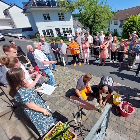Zwei Männer verlegen kniend Stolpersteine im Pflaster, eine Gruppe von Menschen beobachtet und fotografiert die Aktion.