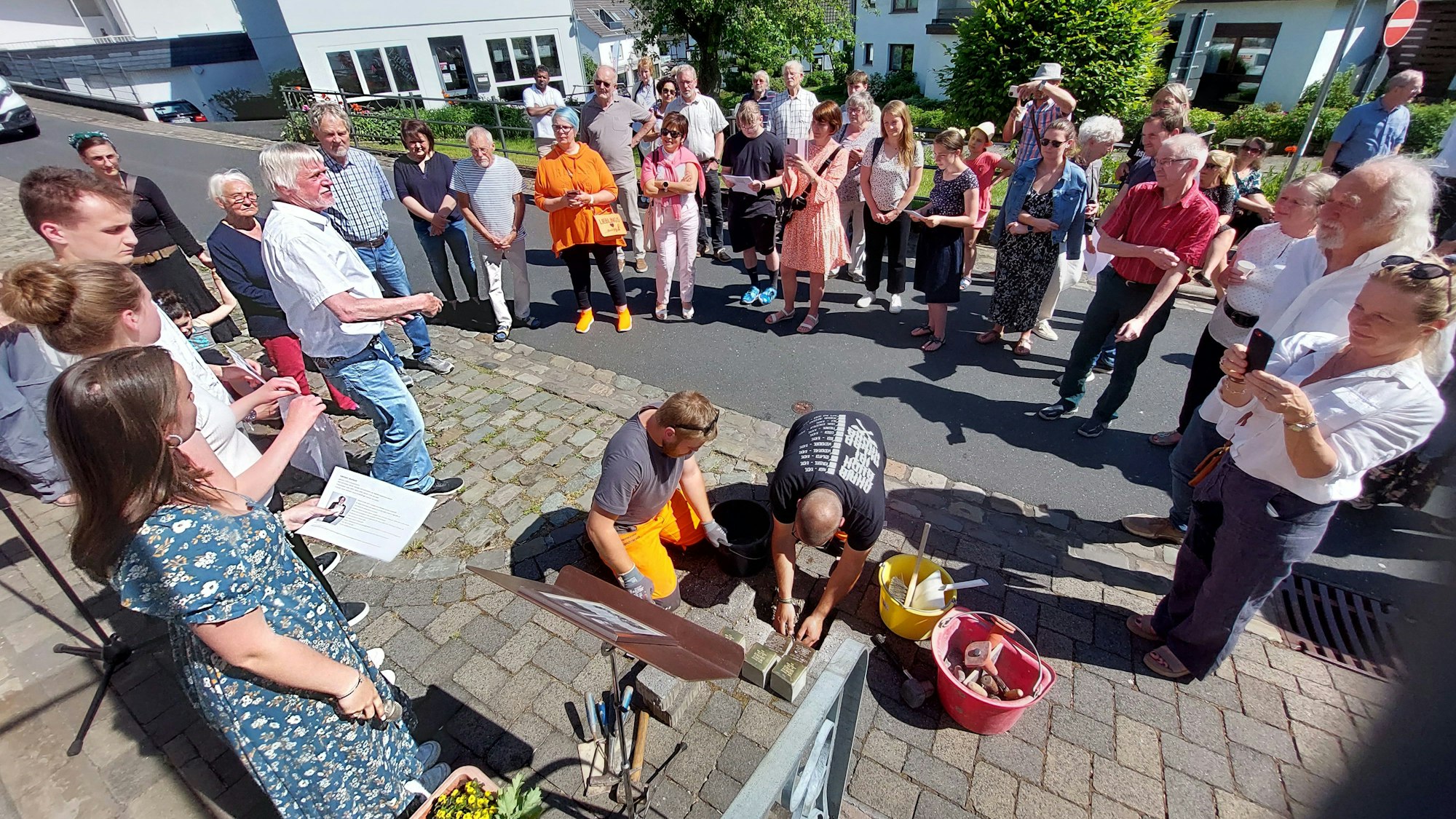 Zwei Männer verlegen kniend Stolpersteine im Pflaster, eine Gruppe von Menschen beobachtet und fotografiert die Aktion.