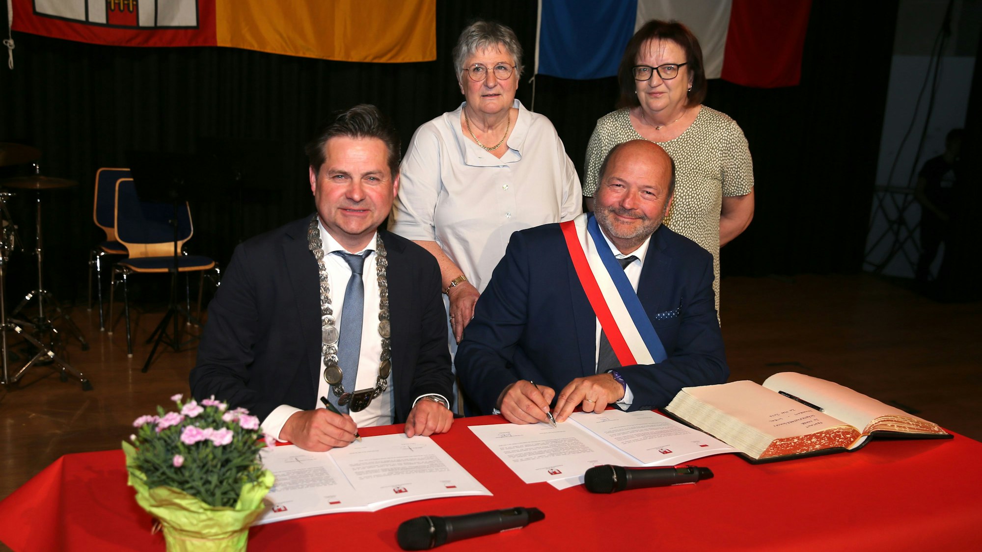 Zwei Bürgermeister sitzen an einem Tisch mit roter Tischdecke und unterzeichnen einen Städtepartnerschaftsvertrag. Daneben liegt das Goldene Buch der Stadt Wiehl. Im Hintergrund stehen zu Frauen.