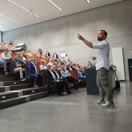 Ein Vortragender steht in einem Hörsaal. Die Sitzreihen sind gefüllt mit Zuhörern.