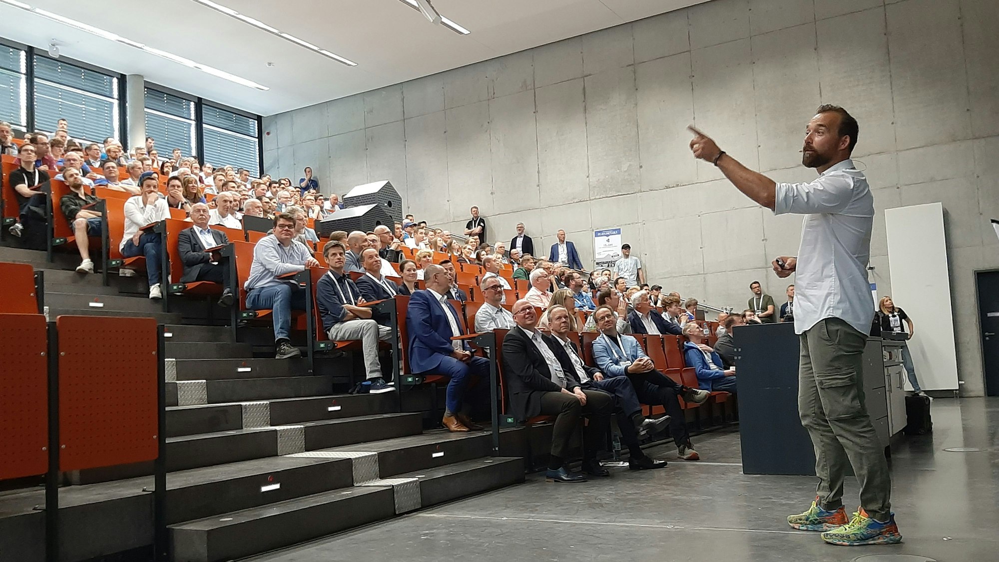 Ein Vortragender steht in einem Hörsaal. Die Sitzreihen sind gefüllt mit Zuhörern.