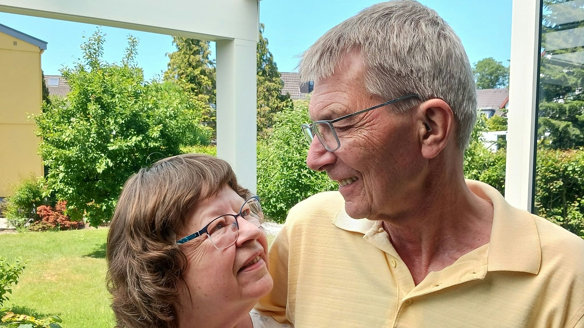 Ein Mann und eine Frau stehen im Garten, halten sich im Arm und sehen sich glücklich lächelnd an.