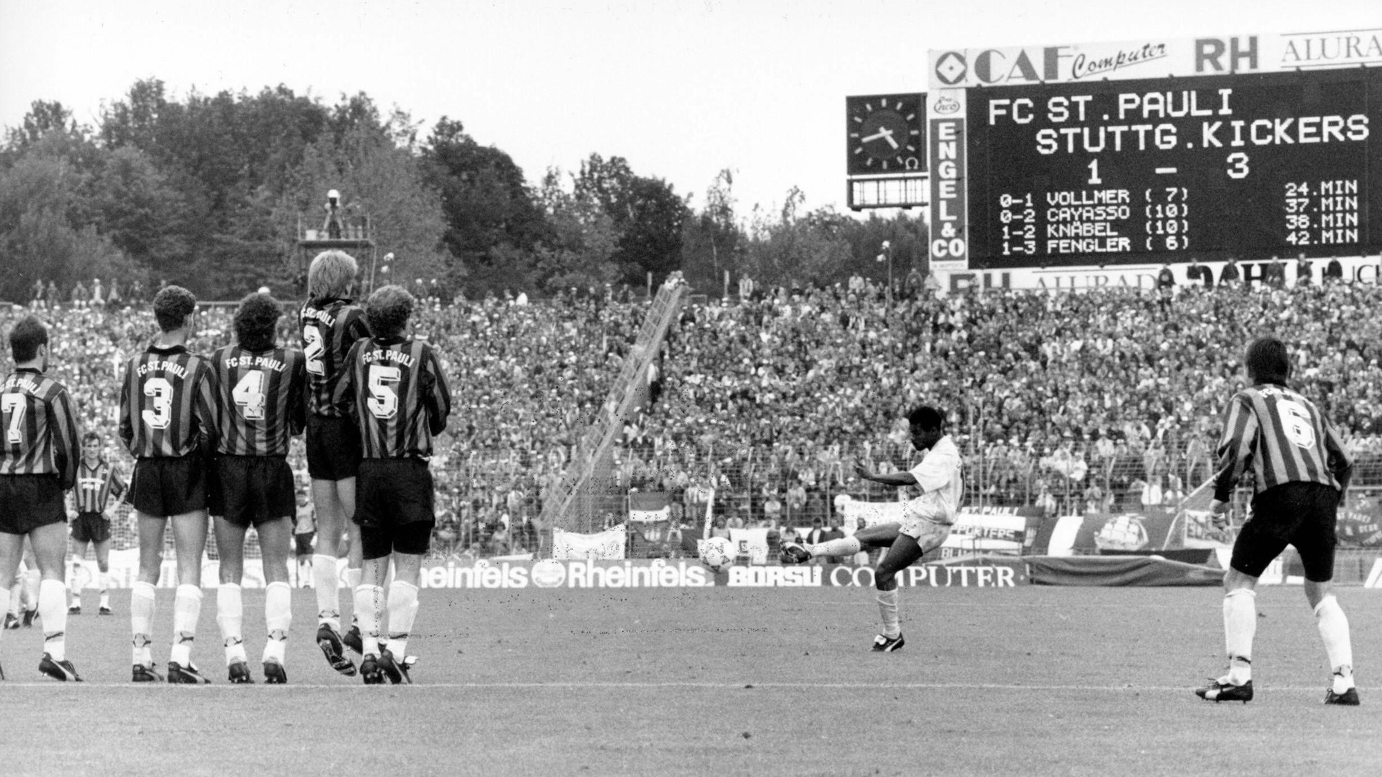 Ein Spieler in heller Kleidung schießt einen Freistoß, während die gegnerischen Spieler eine Mauer bilden. Im Hintergrund zeigt die Anzeigentafel das Ergebnis.
