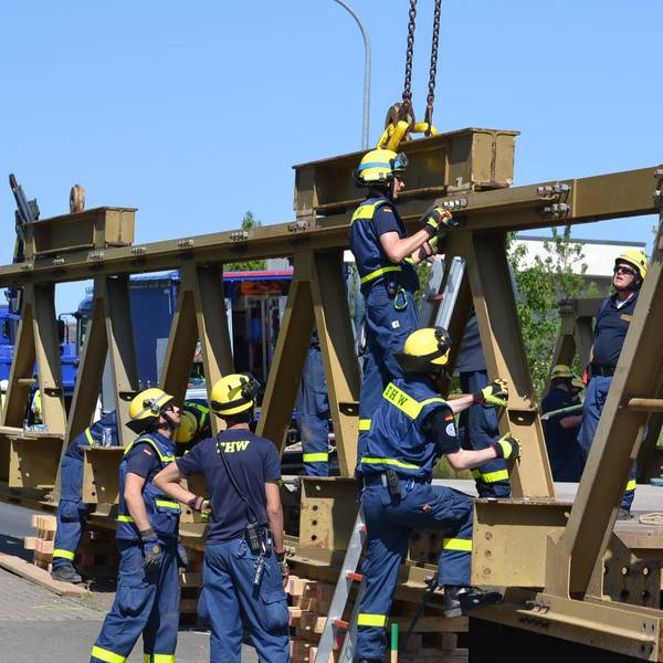 Acht THW-Helfer in blauer Arbeitskleidung und mit gelben Helmen arbeiten am Abbau einer massiven Stahlbrücke.