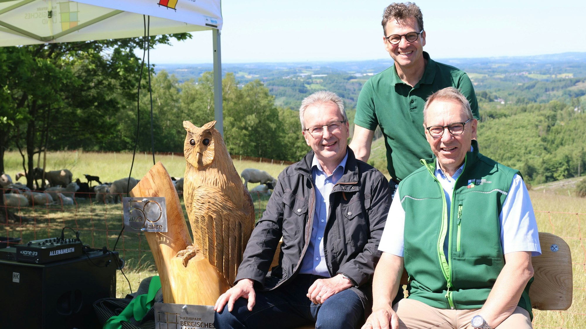 Drei Männer sitzen und stehen an einer Bank aus Holz, an deren Seite eine geschnitzte Eule zu sehen ist.