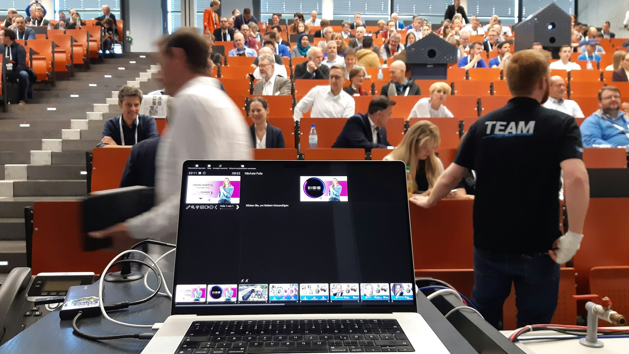 Ein Laptop steht aufgeklappt auf einem Rednerpult. Im Hintergrund ist ein gut gefüllter Vorlesungssaal zu sehen.