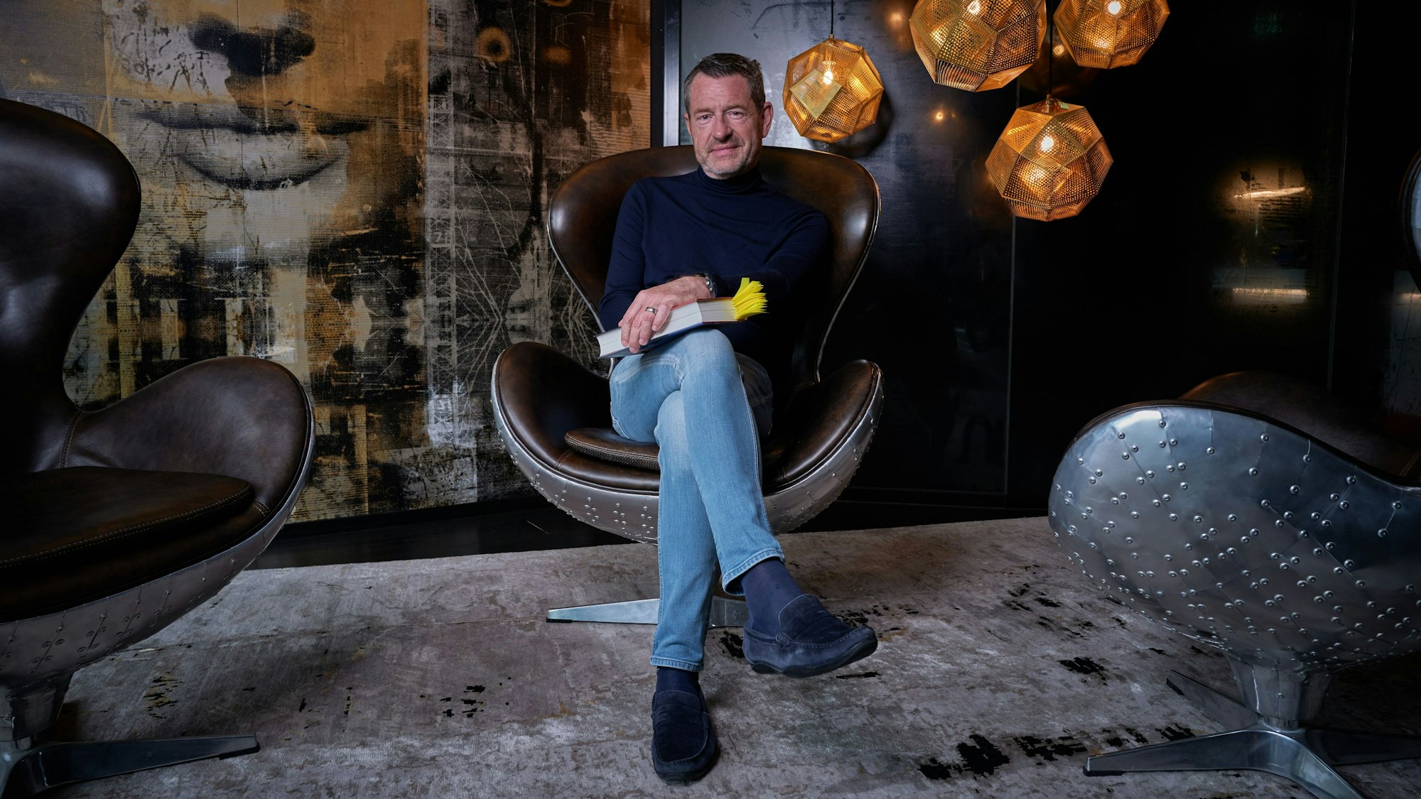 Kai Diekmann sitzt mit überkreuzten Beinen in einem Sessel, er trägt Jeans und einen blauen Rollkragenpullover und hält sein Buch in den Händen.