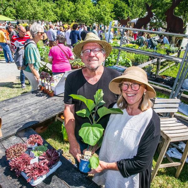 Ein Mann und eine Frau mit Hüten zeigen fröhlich eine Topfpflanze an einem Stand.