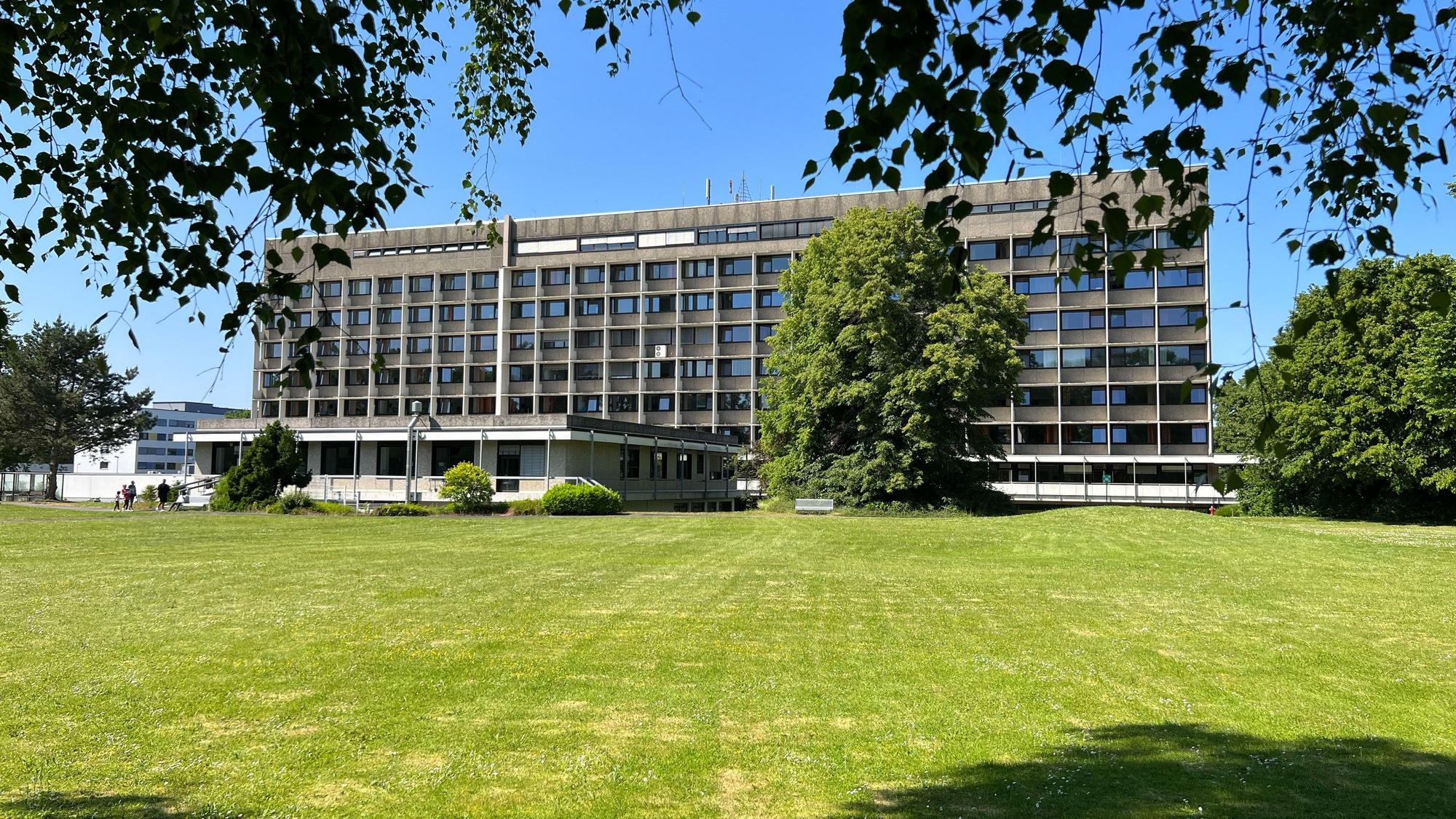 Zu sehen ist die Rückseite des Krankenhauses Porz. Davor steht ein großer grüner Baum und liegt eine grüne Wiese.