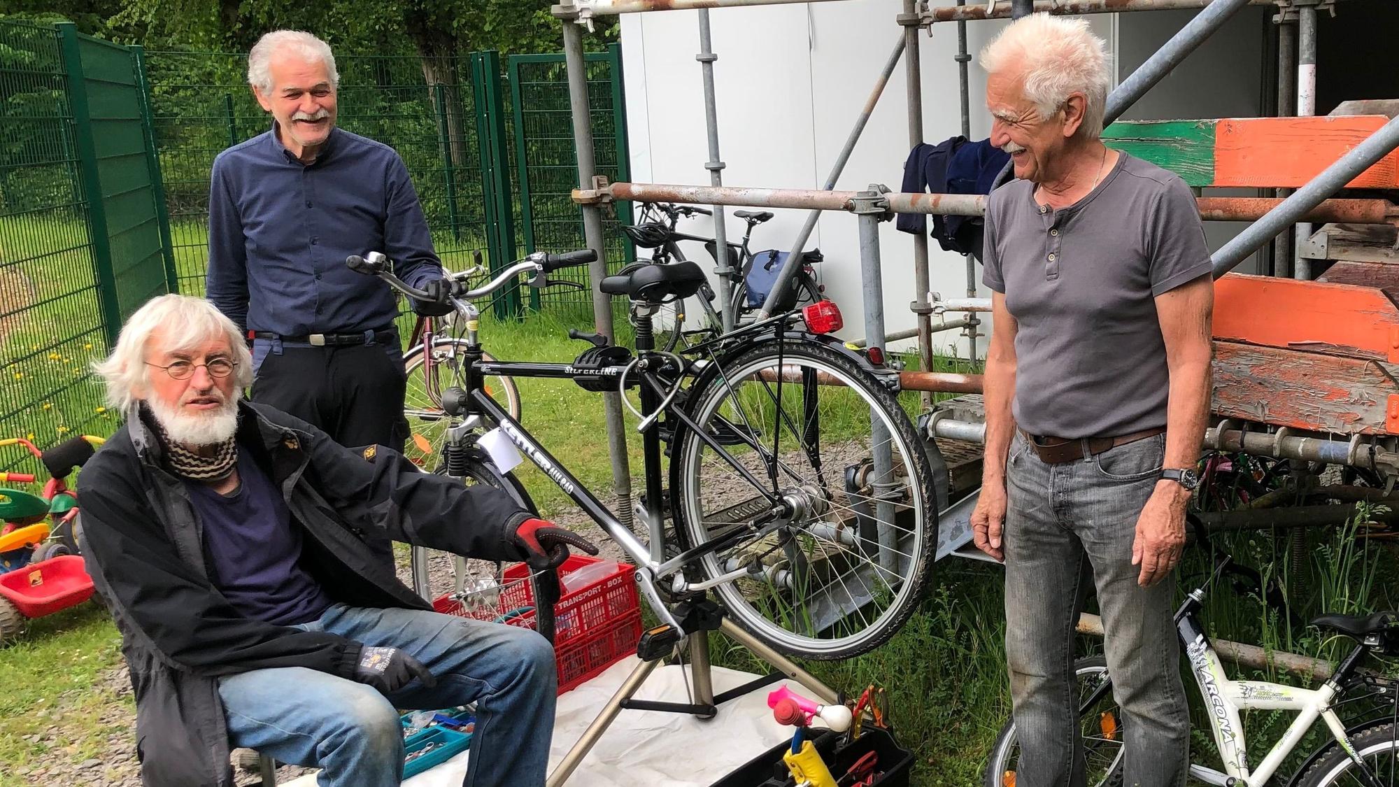 Drei ältere Männer stehen bei einem Fahrrad, das repariert wird, an einem Gerüst.